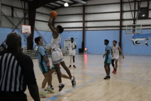 A basketball player leaps to take a shot while teammates and opponents watch in a gymnasium, with a scoreboard visible in the background