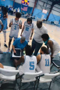 A basketball team in white jerseys gathers around a coach, studying a strategy board during a timeout on the court