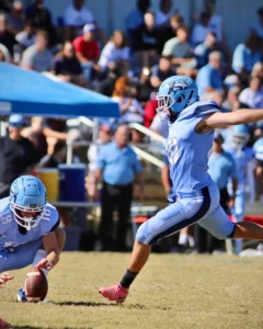 A football player prepares to kick the ball, while a teammate holds it on the field, surrounded by spectators