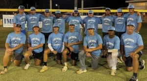 A group of 12 baseball players in blue "Lighthouse Baseball" shirts poses for a team photo on a field at night.