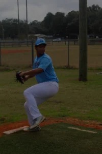 A player wearing a light blue jersey and white pants is preparing to pitch, positioned on a grassy baseball field